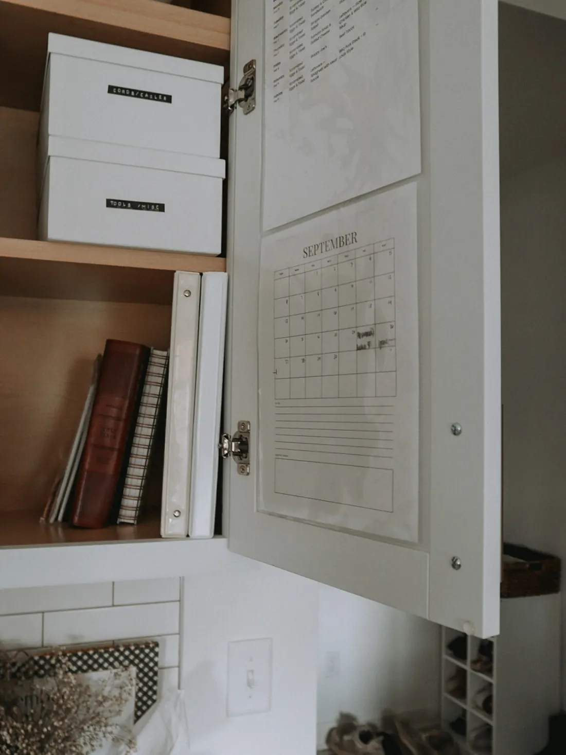 Meal plan and monthly calendar tucked inside an open kitchen cabinet door beside labeled bins and binders